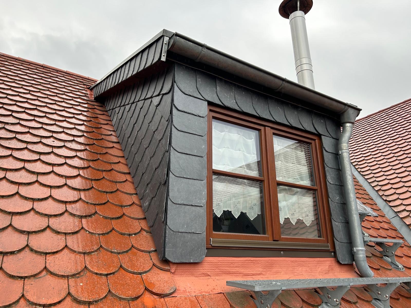 Dach z czerwonej dachówki ceramicznej i lukarna kryta szarym łupkiem, widoczny komin i okno z firankami.