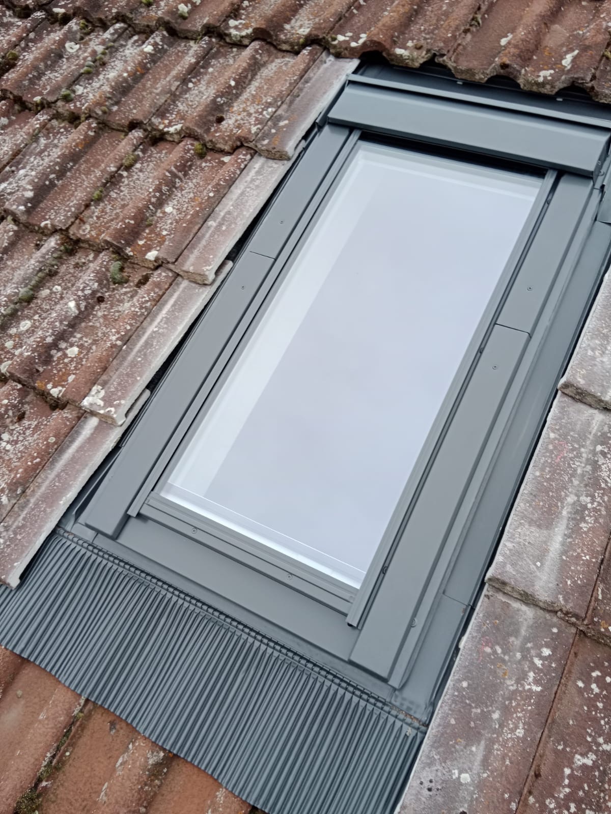 Nowo zamontowane okno dachowe z szarą ramą, wkomponowane w starą, brązową dachówkę pokrytą mchem i plamami. Widoczny element uszczelniający z blachy falistej.