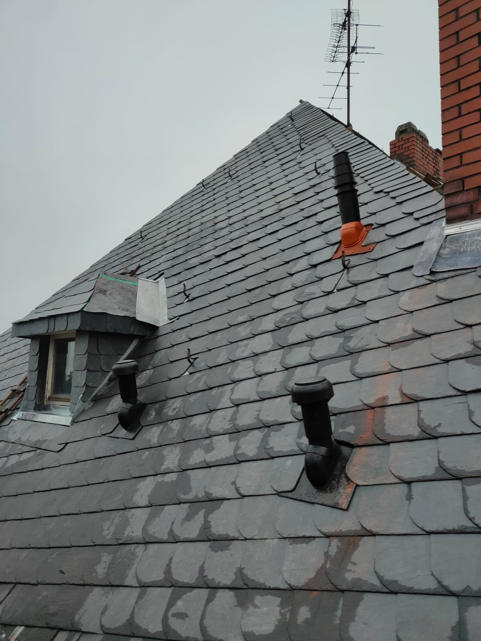 Szary dach kryty łupkiem z oknem dachowym, kominem z czerwonej cegły i anteną telewizyjną na szczycie, widoczne elementy wentylacyjne.