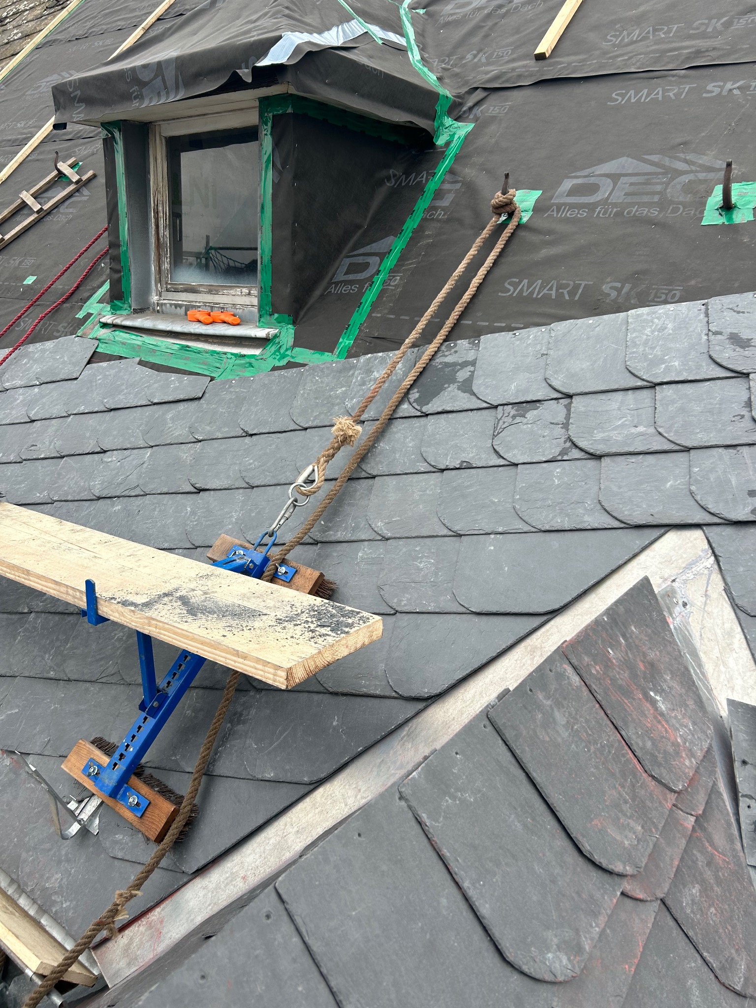 Fragment dachu krytego łupkiem z oknem dachowym w trakcie montażu, widoczna membrana dachowa, lina zabezpieczająca i rusztowanie dekarskie.