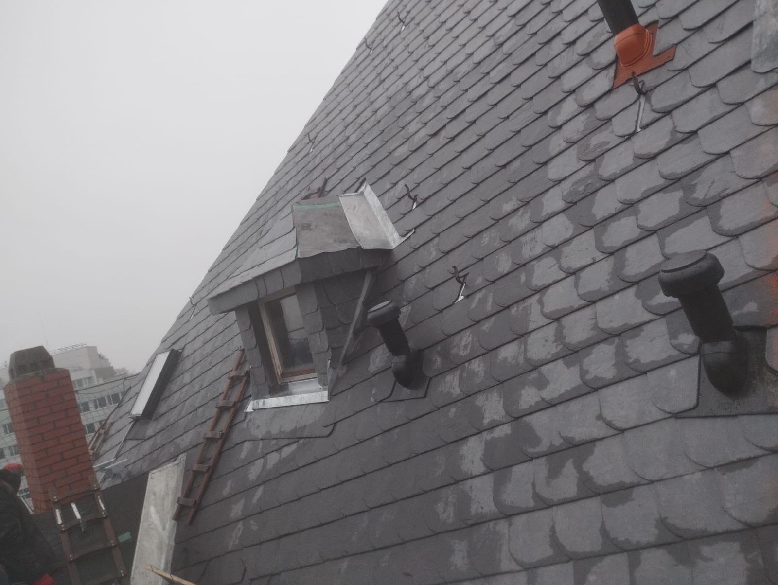 Szary dach pokryty łupkiem z lukarną i kominami wentylacyjnymi, widoczna drabina oparta o dach.