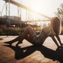 Kobieta w kwiecistej sukience relaksuje się na tle konstrukcji mostu w ciepłym, słonecznym świetle. Delikatny uśmiech i uniesiona głowa dodają uroku.