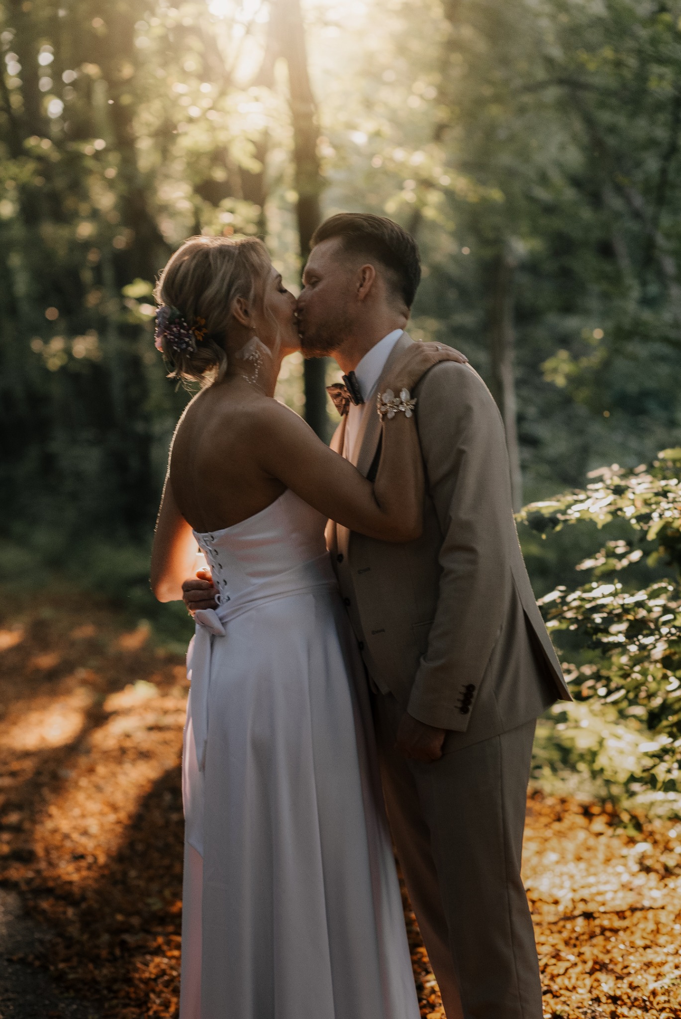 Para młoda w objęciach, pocałunek na tle leśnej scenerii w promieniach zachodzącego słońca. Suknia z gorsetem, muszka i brązowy garnitur.