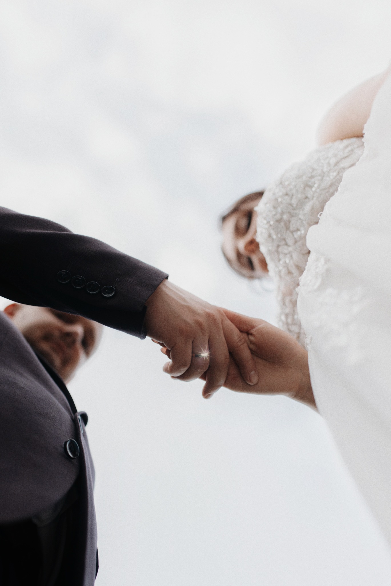 Ujęcie z dołu: panna młoda w sukni i pan młody w garniturze trzymają się za ręce, na palcu obrączka. Delikatne światło podkreśla detale.