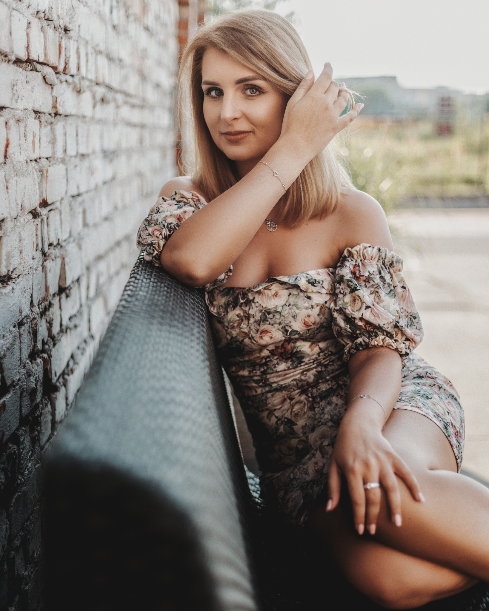 Portret kobiety w kwiecistej sukience siedzącej na ławce, opierającej się o ceglaną ścianę. Subtelna biżuteria, naturalny makijaż, miękkie światło.