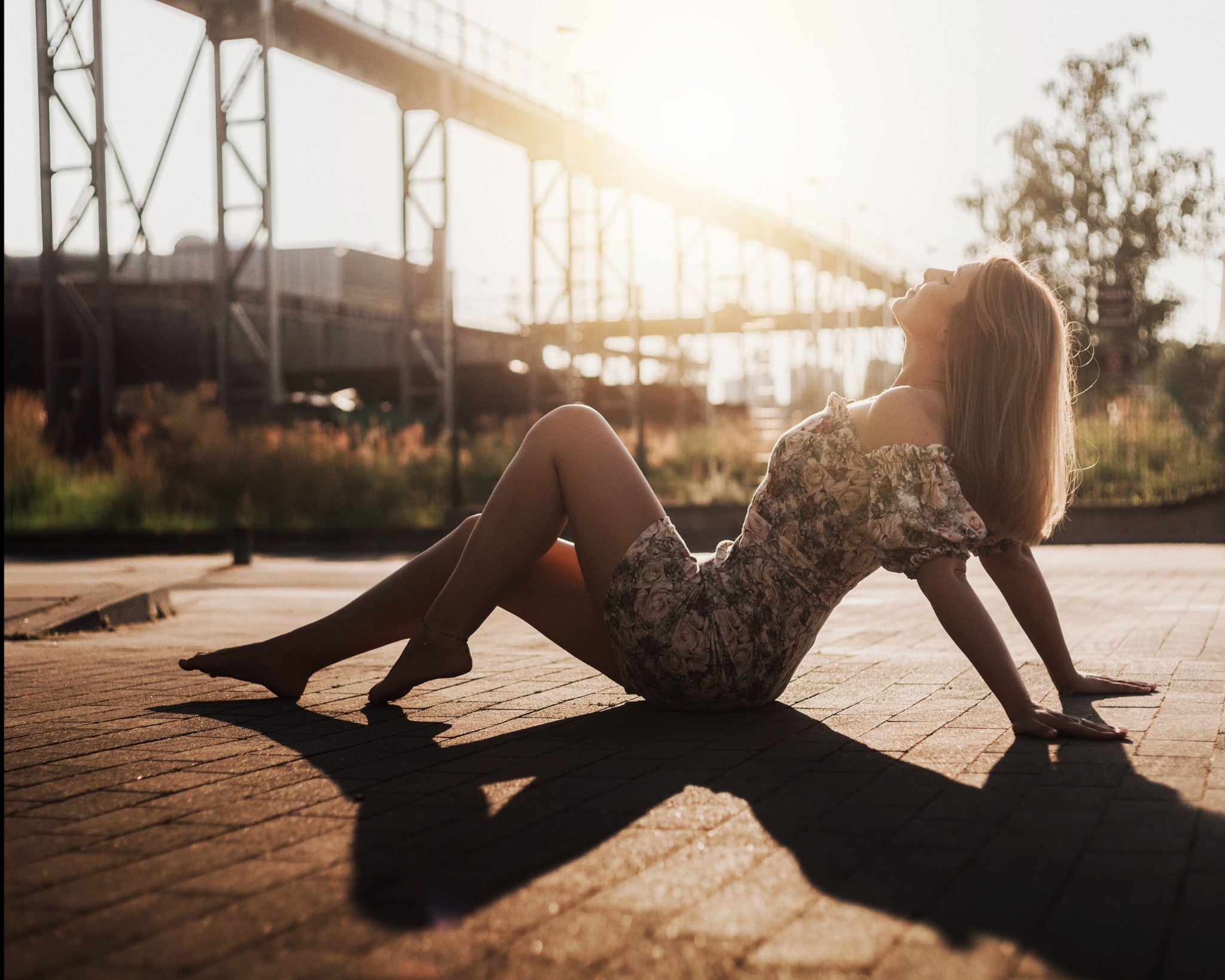 Kobieta w kwiecistej sukience relaksuje się na tle konstrukcji mostu w ciepłym, słonecznym świetle. Delikatny uśmiech i uniesiona głowa dodają uroku.