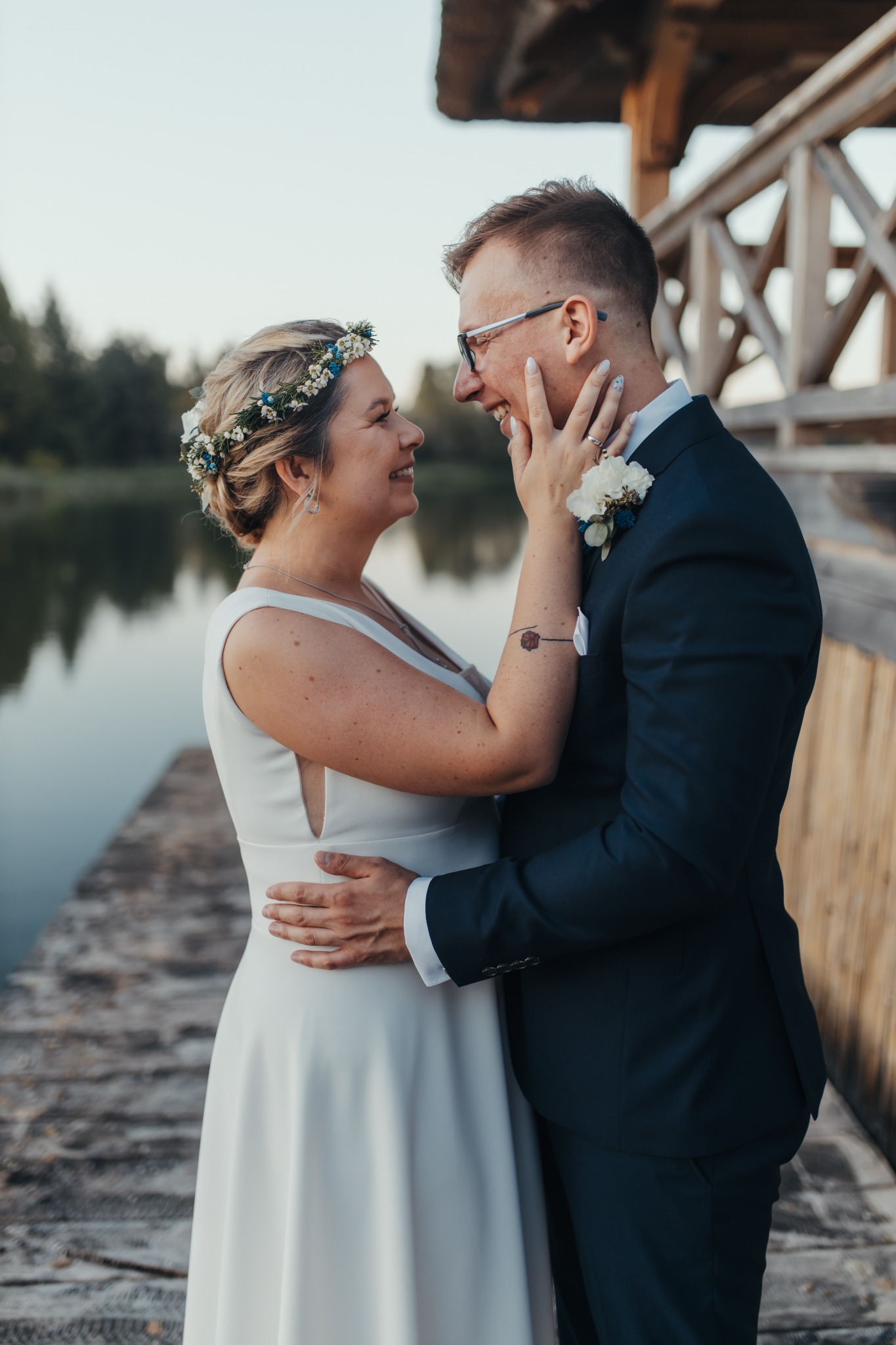 Uśmiechnięta para młoda na drewnianym pomoście nad jeziorem, panna młoda z wiankiem z białych i niebieskich kwiatów, pan młody w granatowym garniturze z białą butonierką i okularach, wzajemnie...