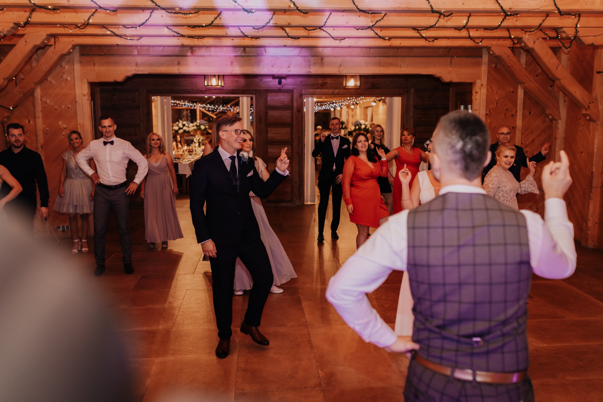 Goście weselni tańczą w drewnianej sali z oświetleniem dekoracyjnym, widoczny pan młody w ciemnym garniturze i okularach, wskazujący palcem w górę.