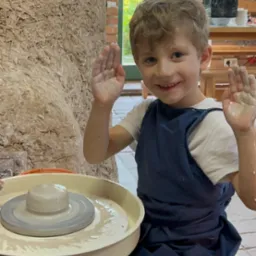 Uśmiechnięty chłopiec w niebieskim fartuchu, z uniesionymi, ubłoconymi rękami, siedzi przy kole garncarskim z glinianym naczyniem w jasnym pomieszczeniu.