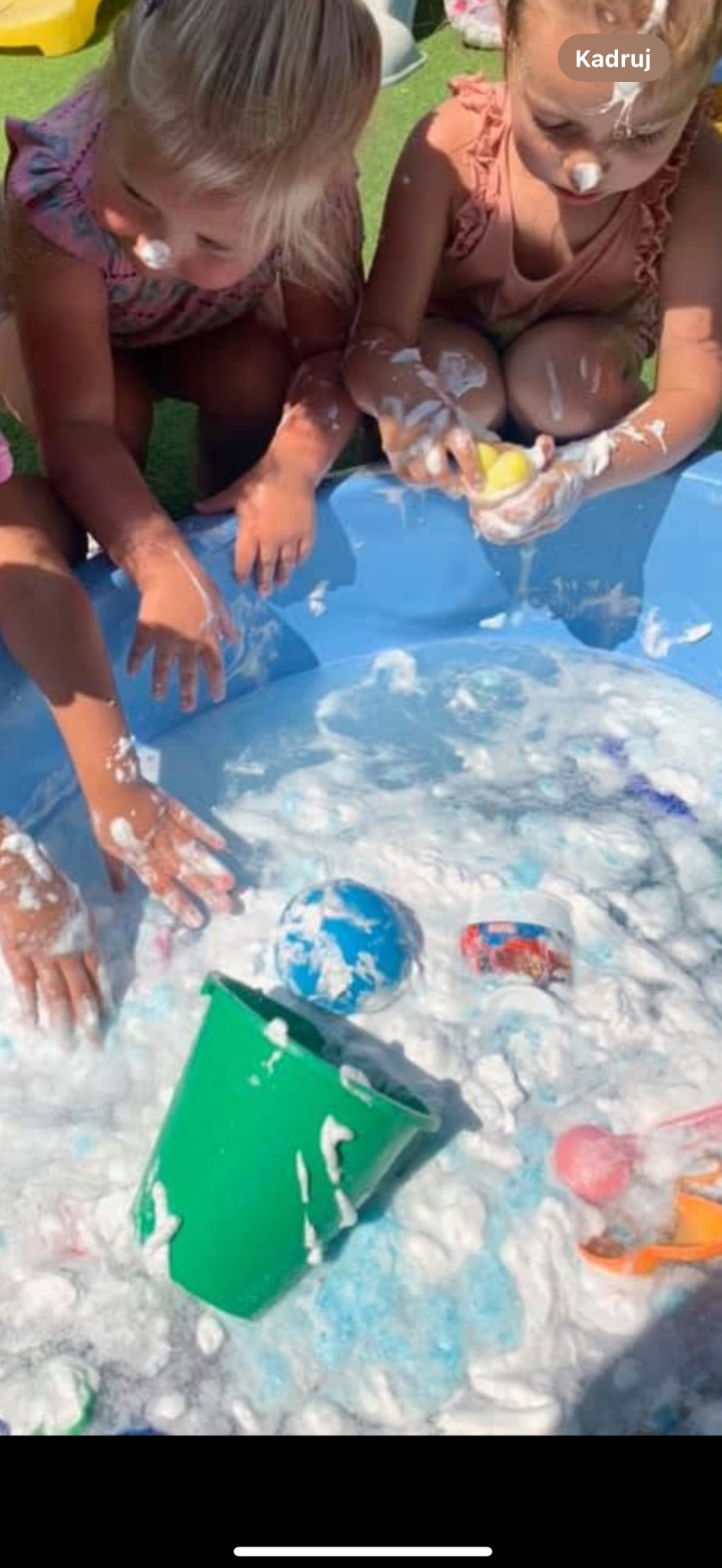 Dwie dziewczynki bawią się w basenie z pianą, mają pianę na nosach. Widoczne zabawki: piłka, wiaderko i inne akcesoria do zabawy w wodzie.