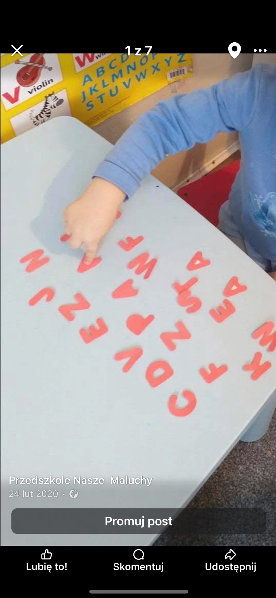 Dziecko układa litery na stole w żłobku. Na stole rozłożone czerwone litery alfabetu. W tle plakat z alfabetem i obrazkami.