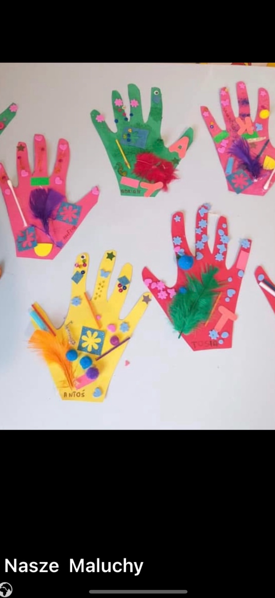 Ręcznie robione, kolorowe prace plastyczne w kształcie dłoni, ozdobione piórami, cekinami i papierowymi elementami, przyklejone na ścianie. Dziecięce imiona na każdej pracy.