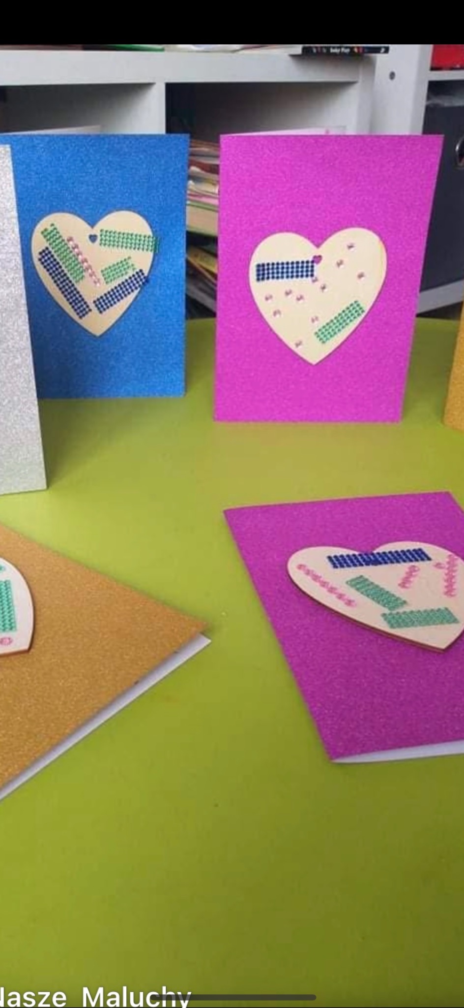 Ręcznie robione kartki z brokatowego papieru w kolorach: niebieskim, różowym, złotym. Na każdej kartce naklejone serce ozdobione kolorową taśmą washi. Prace dzieci w żłobku.