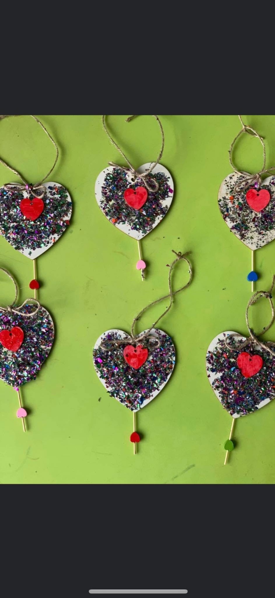 Sześć ręcznie robionych ozdób w kształcie serca z brokatem i drewnianymi elementami na zielonym tle, zawieszone na sznurkach. Elementy dekoracyjne wykonane przez dzieci.