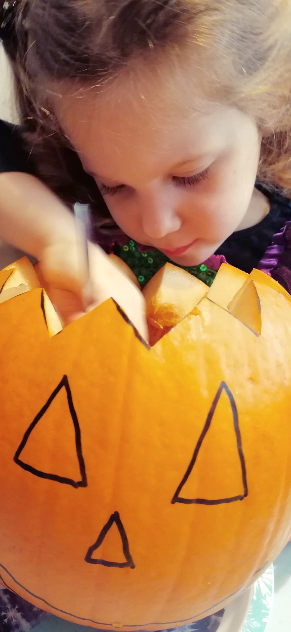Dziewczynka w kostiumie wyjmuje miąższ z wydrążonej dyni z narysowanymi trójkątami, przygotowując dekorację na Halloween.