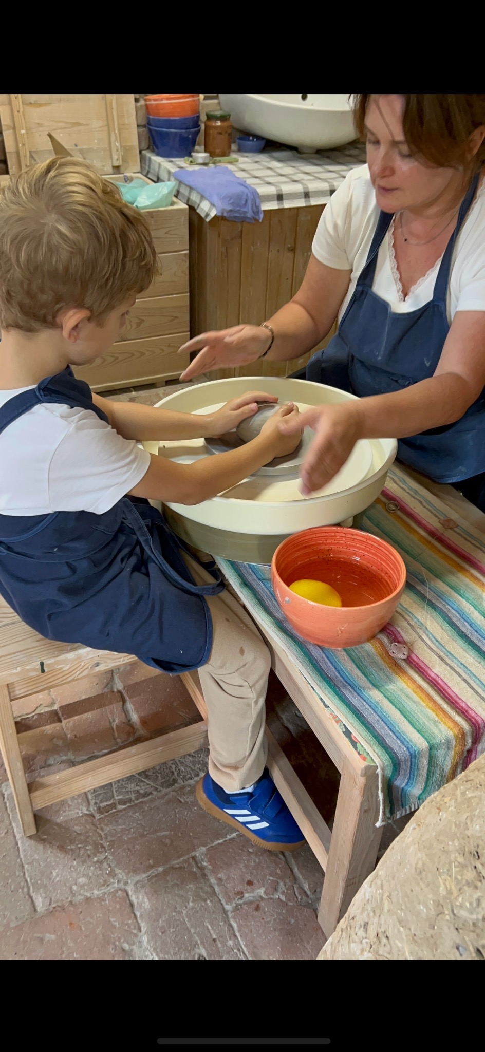 Chłopiec w niebieskim fartuchu uczy się toczenia gliny na kole garncarskim pod okiem instruktorki. W tle widoczne ceramiczne naczynia i ceglana podłoga.