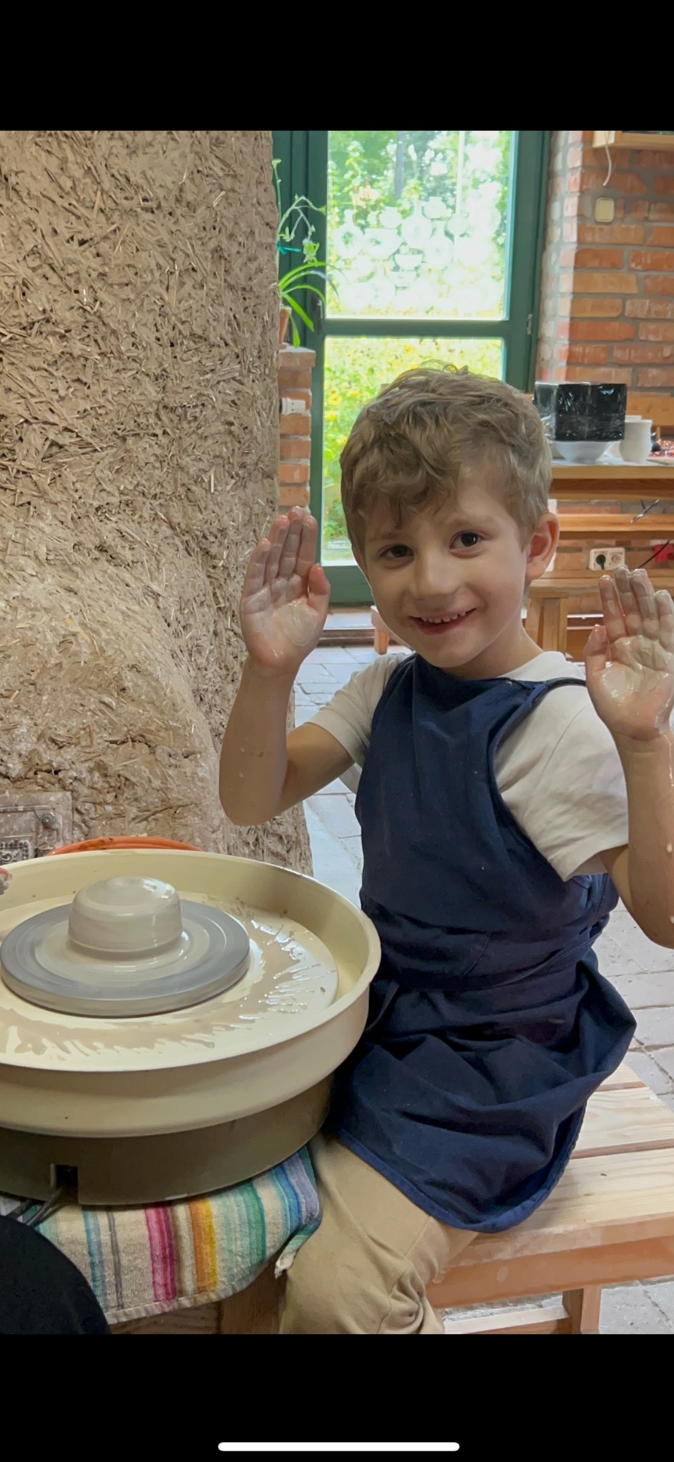 Uśmiechnięty chłopiec w niebieskim fartuchu, z uniesionymi, ubłoconymi rękami, siedzi przy kole garncarskim z glinianym naczyniem w jasnym pomieszczeniu.