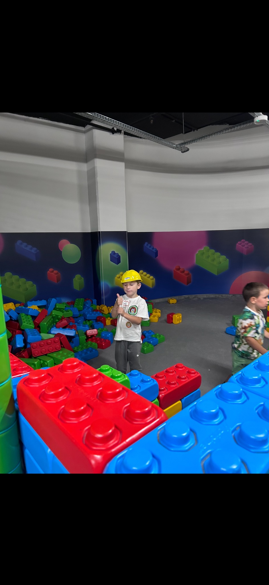 Chłopiec w kasku budowniczego bawi się klockami w sali zabaw na tle kolorowych dekoracji, pokazując kciuk w górę.