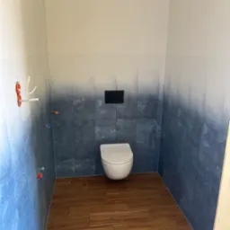 Nowoczesna toaleta w trakcie remontu: biała miska wisząca, ściany wyłożone gradientowymi płytkami w odcieniach bieli i granatu, imitacja drewnianej podłogi z płytek, widoczne wystające kable...