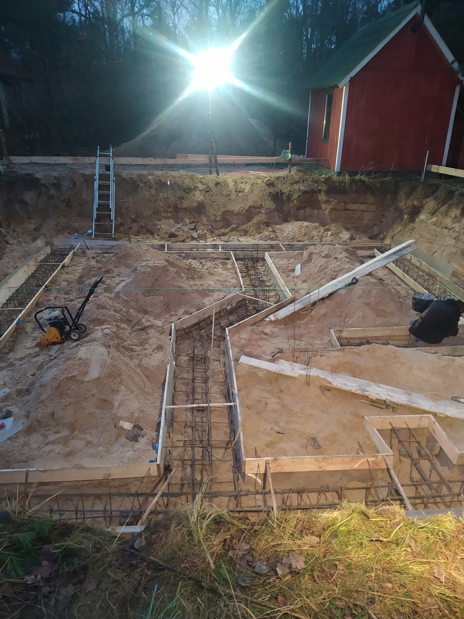 Przygotowanie fundamentów pod budynek: wykop, szalunki zbrojeniowe wypełnione piaskiem, ubijarka wibracyjna, drabina oparta o skarpę, w tle czerwony budynek i drzewa, silne światło słoneczne.