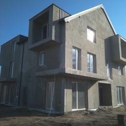 Surowy, dwupiętrowy dom w stanie deweloperskim, z widoczną elewacją w szarym tynku i dużymi oknami, na tle błękitnego nieba.