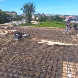 Zbrojenie płyty fundamentowej domu jednorodzinnego, widok z góry, dwóch pracowników w tle podczas pracy, miasto w oddali, niebieskie niebo.