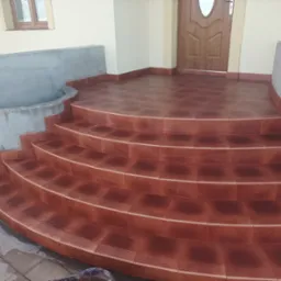 Wykończone schody wejściowe do domu, wyłożone brązowymi, kwadratowymi płytkami z delikatnym wzorem, z widocznym fragmentem elewacji i stolarki okiennej.