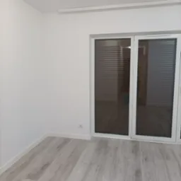 Puste, białe wnętrze z panelami podłogowymi imitującymi drewno i dużym oknem balkonowym z żaluzjami.