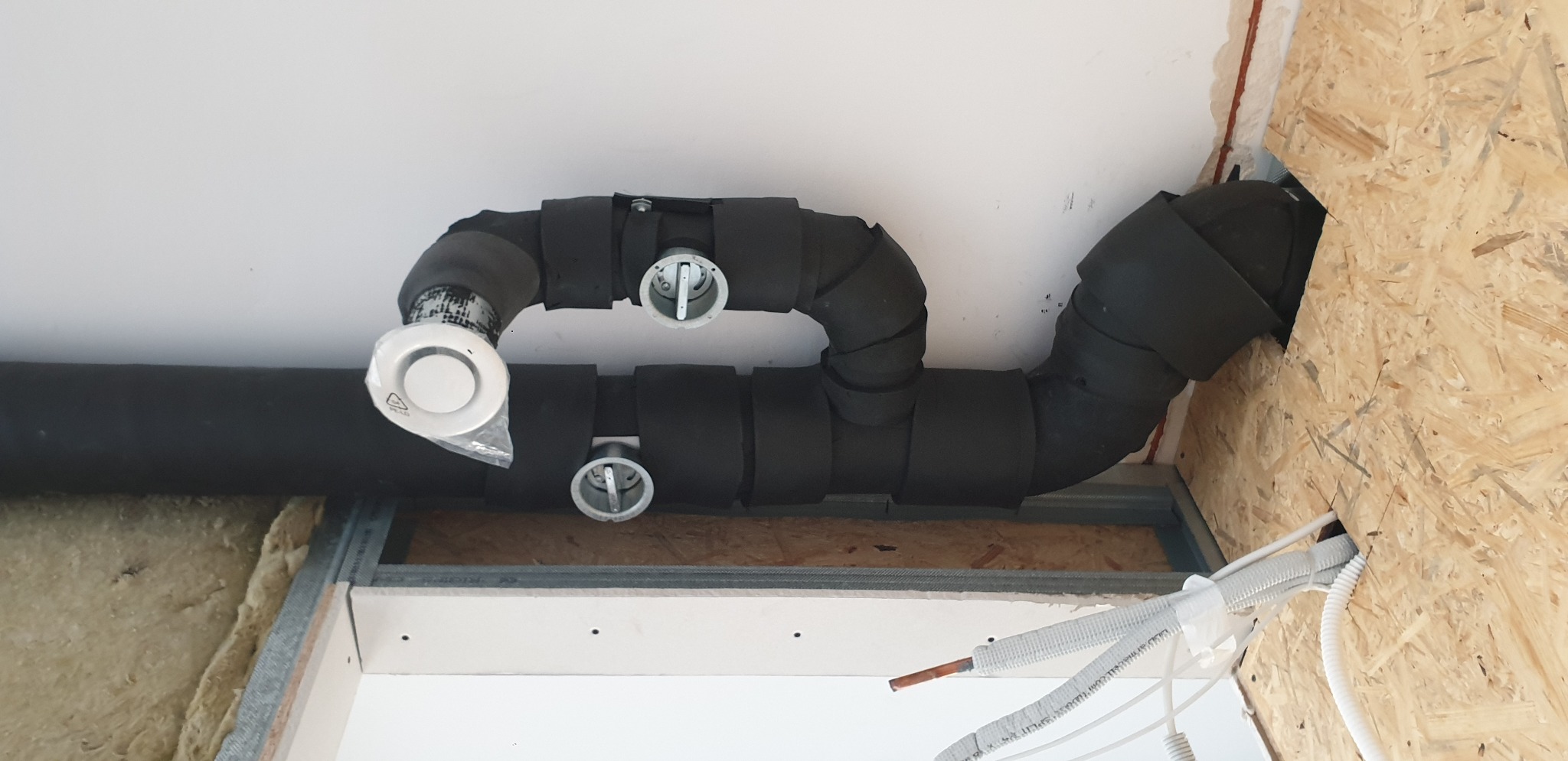 Czarna, zaizolowana rura wentylacyjna pod sufitem z widocznymi elementami montażowymi i przewodami elektrycznymi w trakcie montażu.