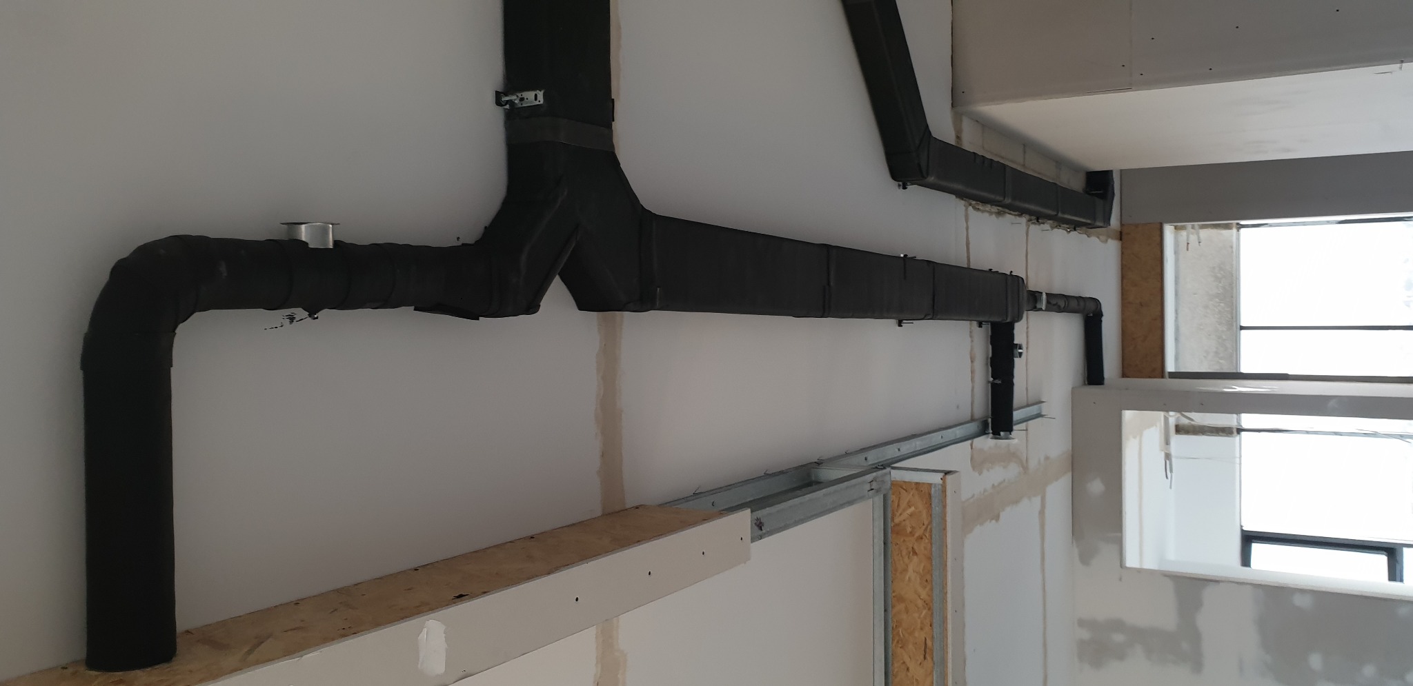Czarne, zaizolowane rury wentylacyjne rozchodzą się w kształcie litery Y na białej ścianie w surowym wnętrzu, z widocznymi elementami konstrukcyjnymi i ramą okienną w tle.
