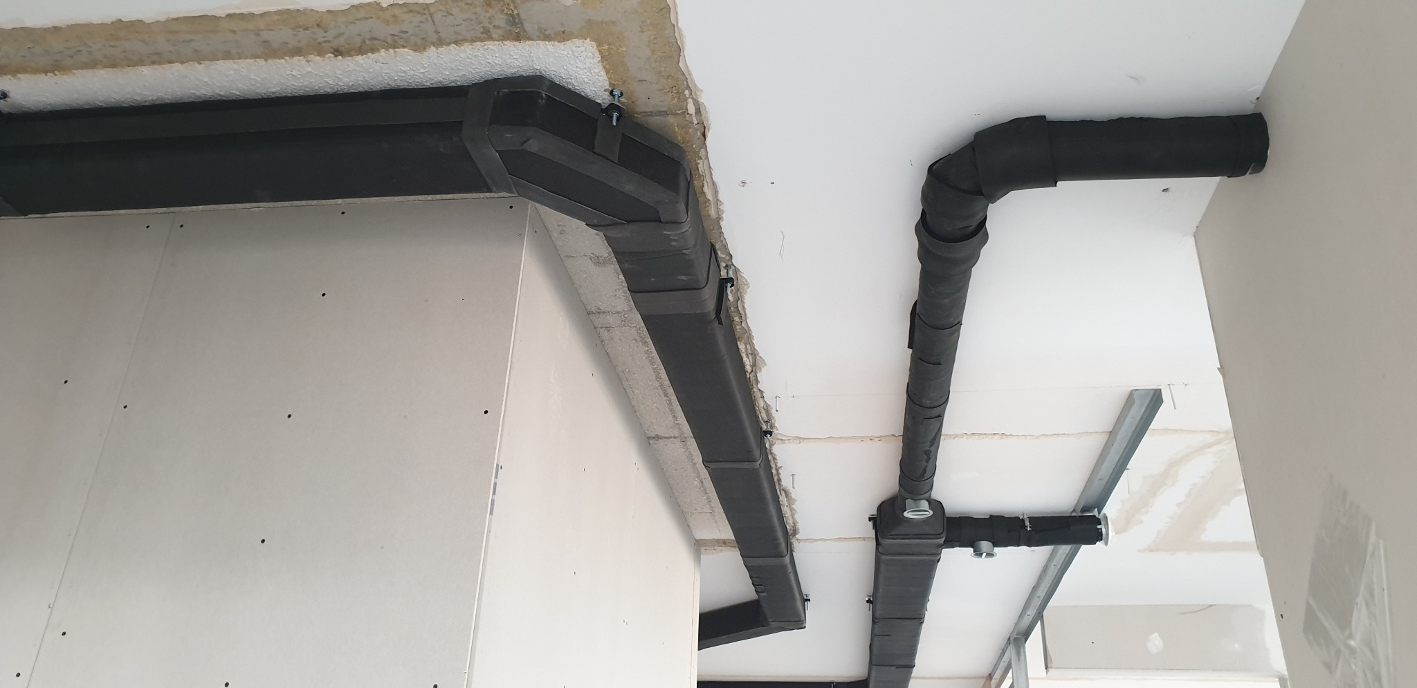 Czarna, izolowana instalacja wentylacyjna pod sufitem w trakcie montażu, widoczne elementy mocujące i surowy stan wykończenia wnętrza.