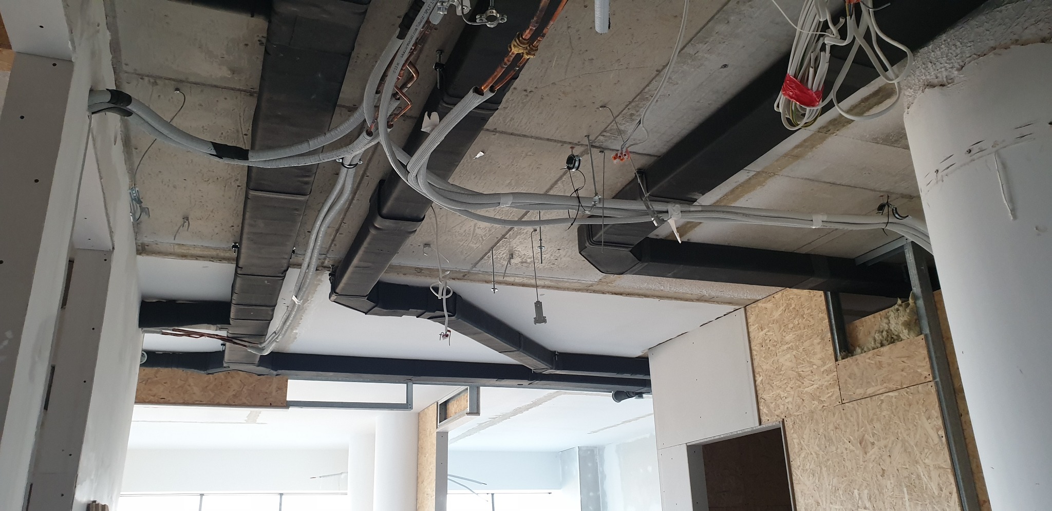 Widok na sufit w trakcie prac wykończeniowych: widoczne kable w peszlach, rury miedziane, czarne kanały wentylacyjne, płyty OSB i fragmenty wełny mineralnej w ścianach działowych.