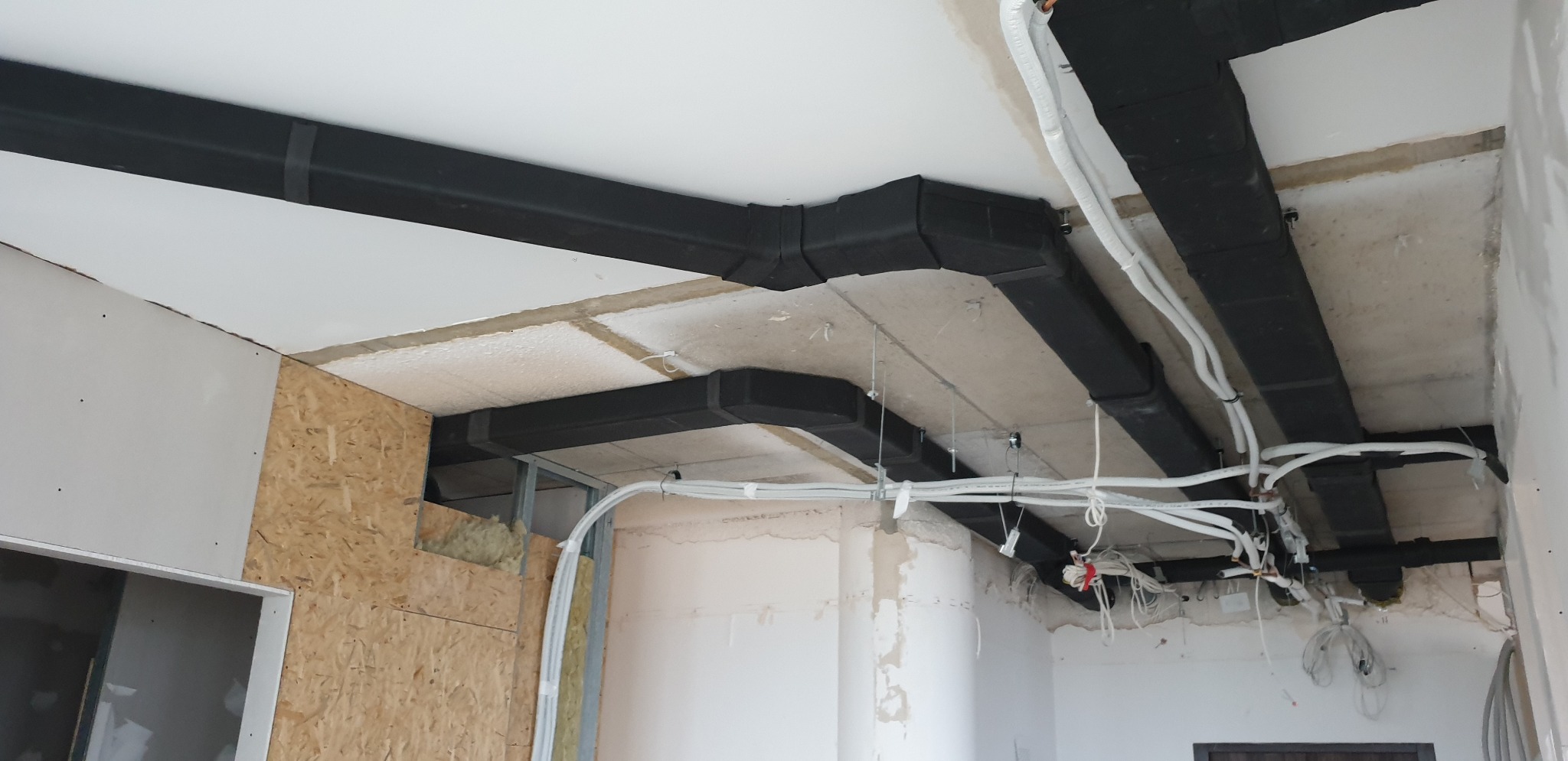 Niedokończone wnętrze z widocznymi czarnymi, zaizolowanymi kanałami wentylacyjnymi pod sufitem oraz przewodami elektrycznymi, ściany w stanie surowym z fragmentami płyt OSB.