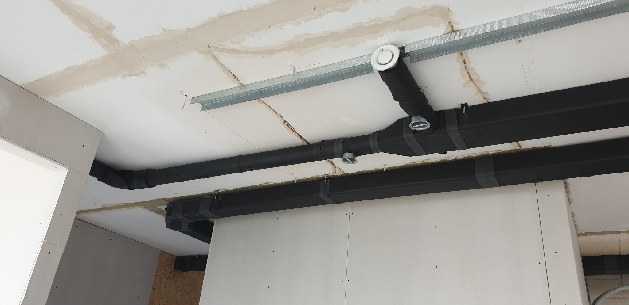 Czarny, zaizolowany system rur wentylacyjnych z okrągłymi dyfuzorami zamontowany pod sufitem podwieszanym w trakcie montażu płyt gipsowo-kartonowych.