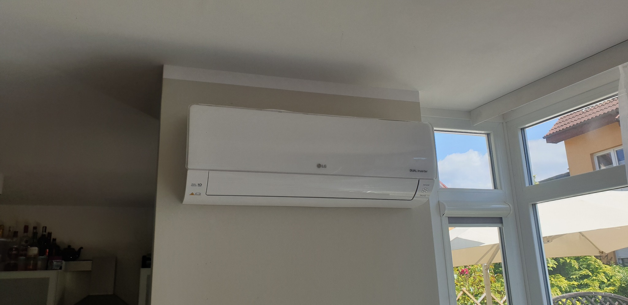 Biały klimatyzator LG Dual Inverter zamontowany na ścianie w jasnym wnętrzu, widok z boku, z oknem i fragmentem ogrodu w tle.