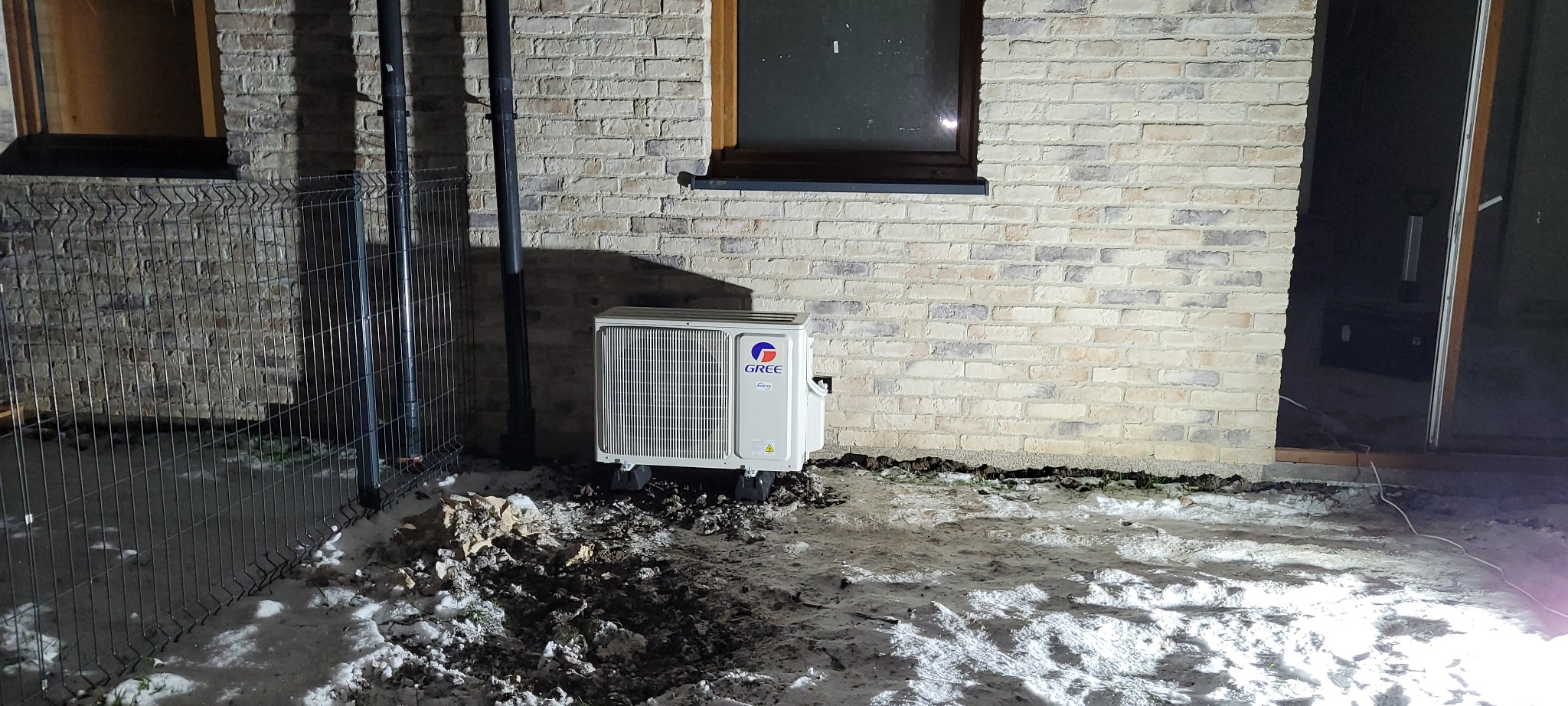 Zewnętrzna jednostka klimatyzacji marki Gree na tymczasowych podstawkach, ustawiona na ubłoconym śniegu przy ścianie budynku z cegły licówki i metalowym ogrodzeniu.