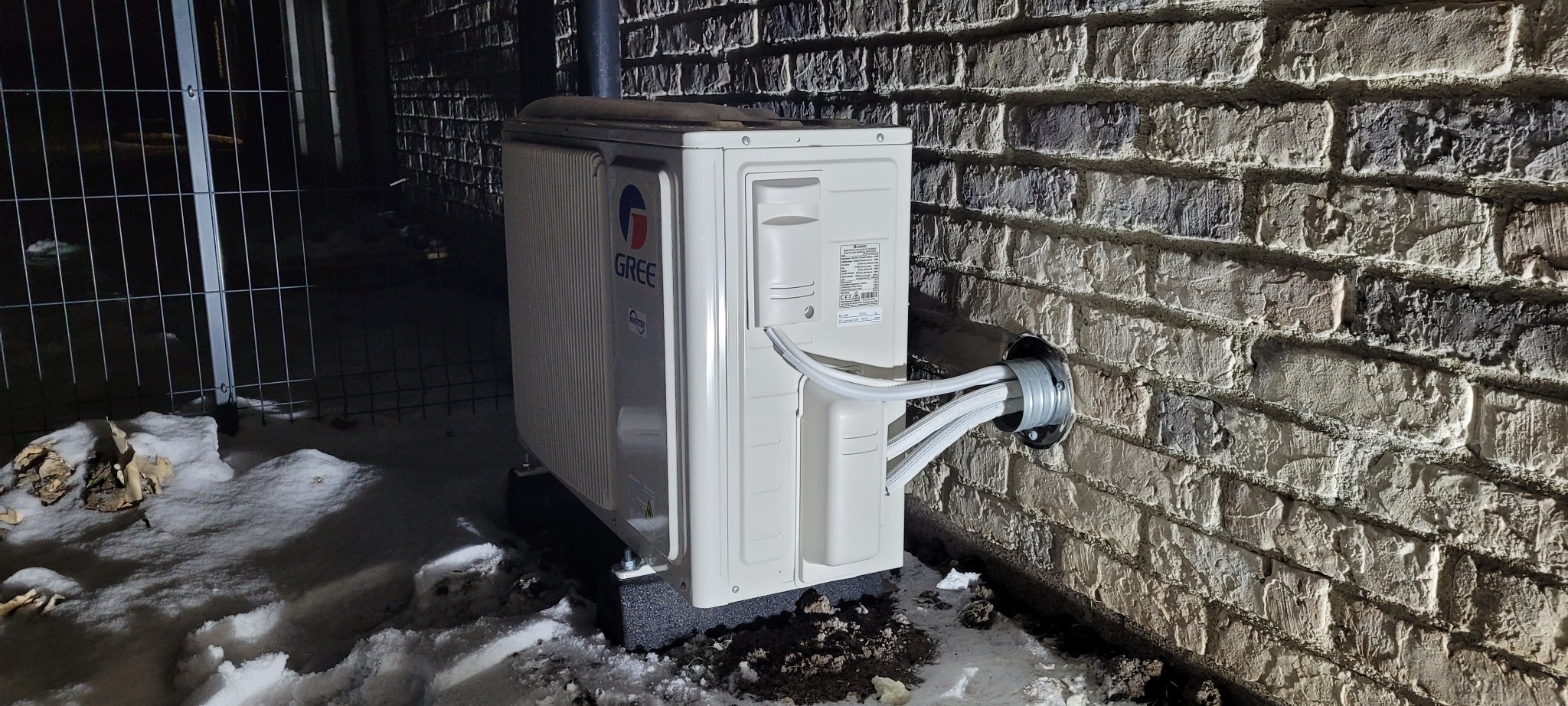 Zewnętrzna jednostka klimatyzacji marki Gree zamontowana na ceglanej ścianie budynku, widoczne przewody w osłonie wchodzące do ściany, śnieg na ziemi.