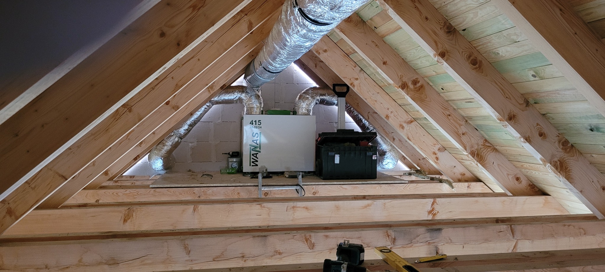Rekuperator WANAS 415 V-TECH z podłączonymi przewodami wentylacyjnymi w przestrzeni poddasza o konstrukcji drewnianej, obok skrzynka narzędziowa.