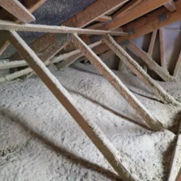 Poddasze z drewnianą konstrukcją krokwi pokryte grubą warstwą jasnego materiału izolacyjnego, prawdopodobnie natryskiwanego, z widocznymi cieniami i nierównomierną strukturą.