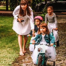 Marta Wójcik - Kobieta w białej sukience i sandałach prowadzi grupę dziewczynek w białych sukienkach na rowerkach po kamiennej ścieżce w ogrodzie.