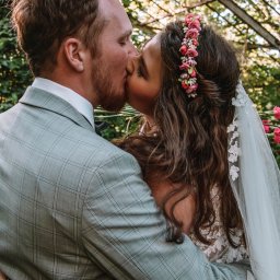 Marta Wójcik - Pocałunek pary młodej pod konstrukcją z pnączy; panna młoda z wiankiem z różowych kwiatów i welonem z aplikacjami, pan młody w szarym garniturze w kratę.