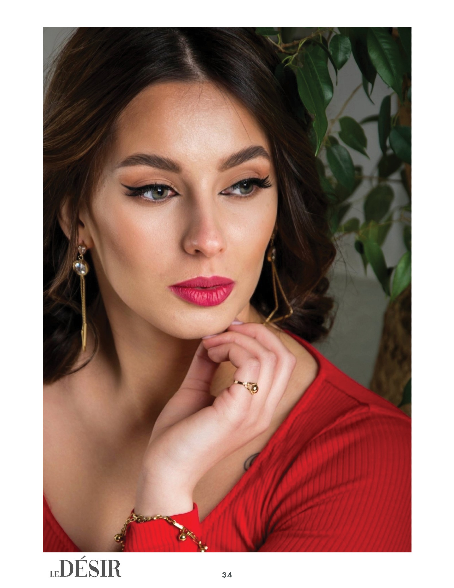 Portret kobiety w czerwonej bluzce, z wyrazistym makijażem, złotą biżuterią, zamyślonym spojrzeniem w bok, na tle zielonych liści.