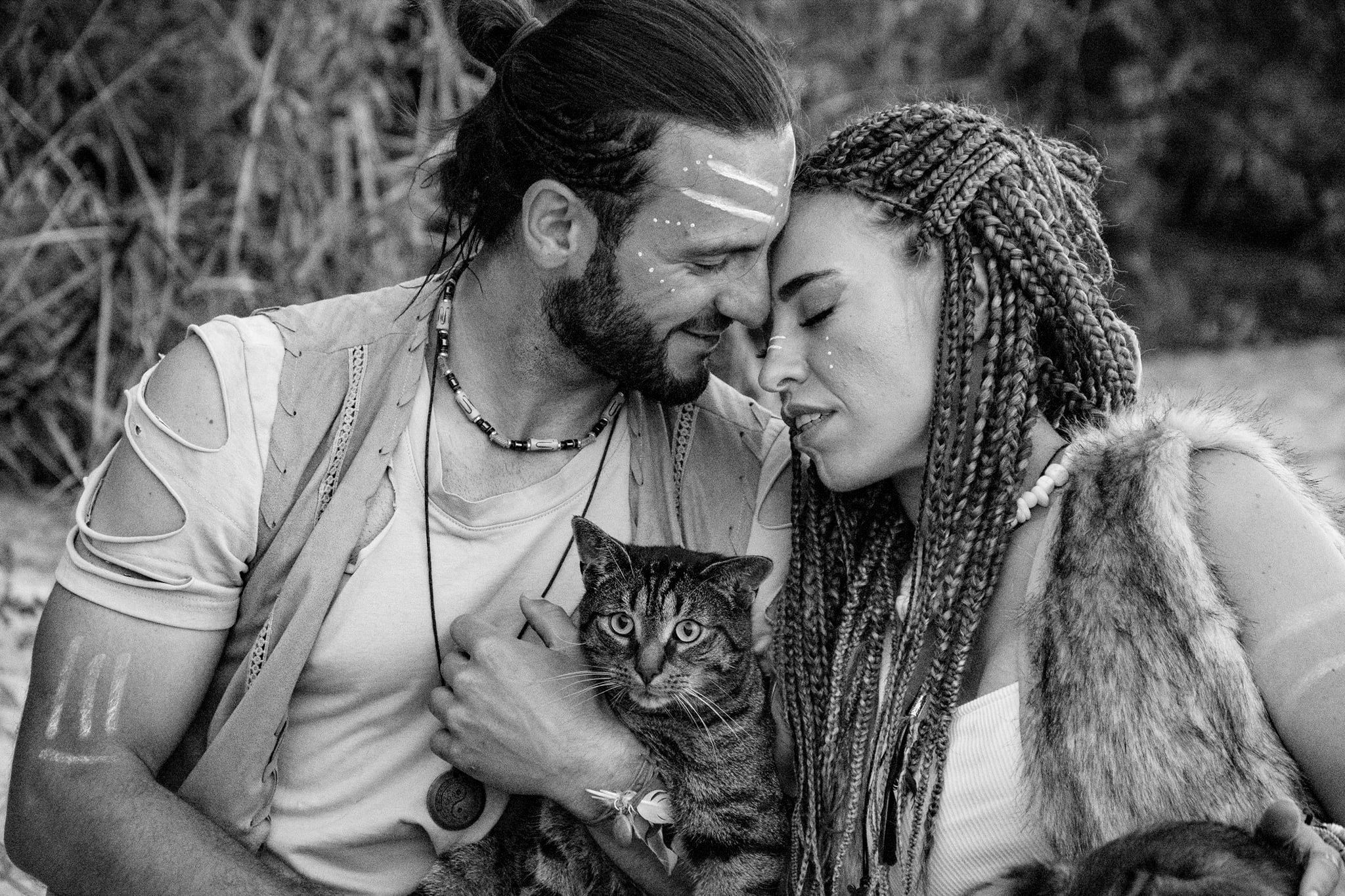 Czarno-białe zdjęcie pary w stylizowanych strojach etnicznych, z pomalowanymi twarzami i kotem na rękach mężczyzny, oboje z zamkniętymi oczami, blisko siebie, w plenerze.