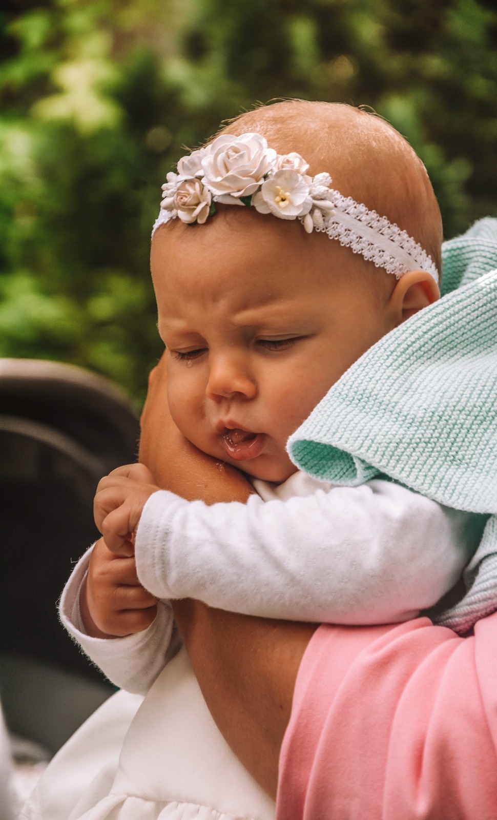 Śpiąca niemowlęca dziewczynka w białej sukience i kwiatowej opasce, trzymana na rękach, z zielonym kocem w tle.