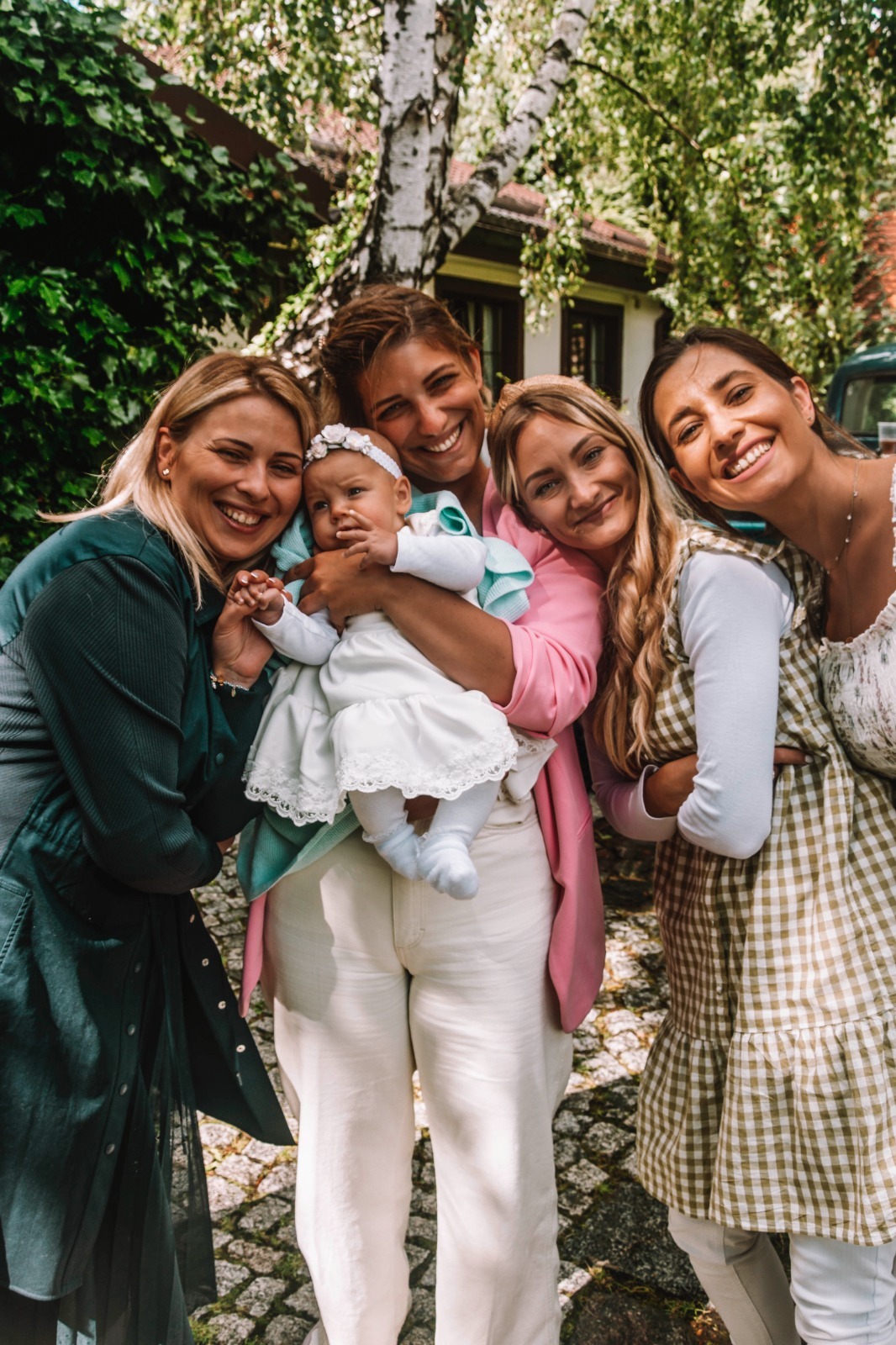 Cztery uśmiechnięte kobiety pozują z niemowlęciem w białej sukience i opasce na głowie, na tle zieleni i białej brzozy.