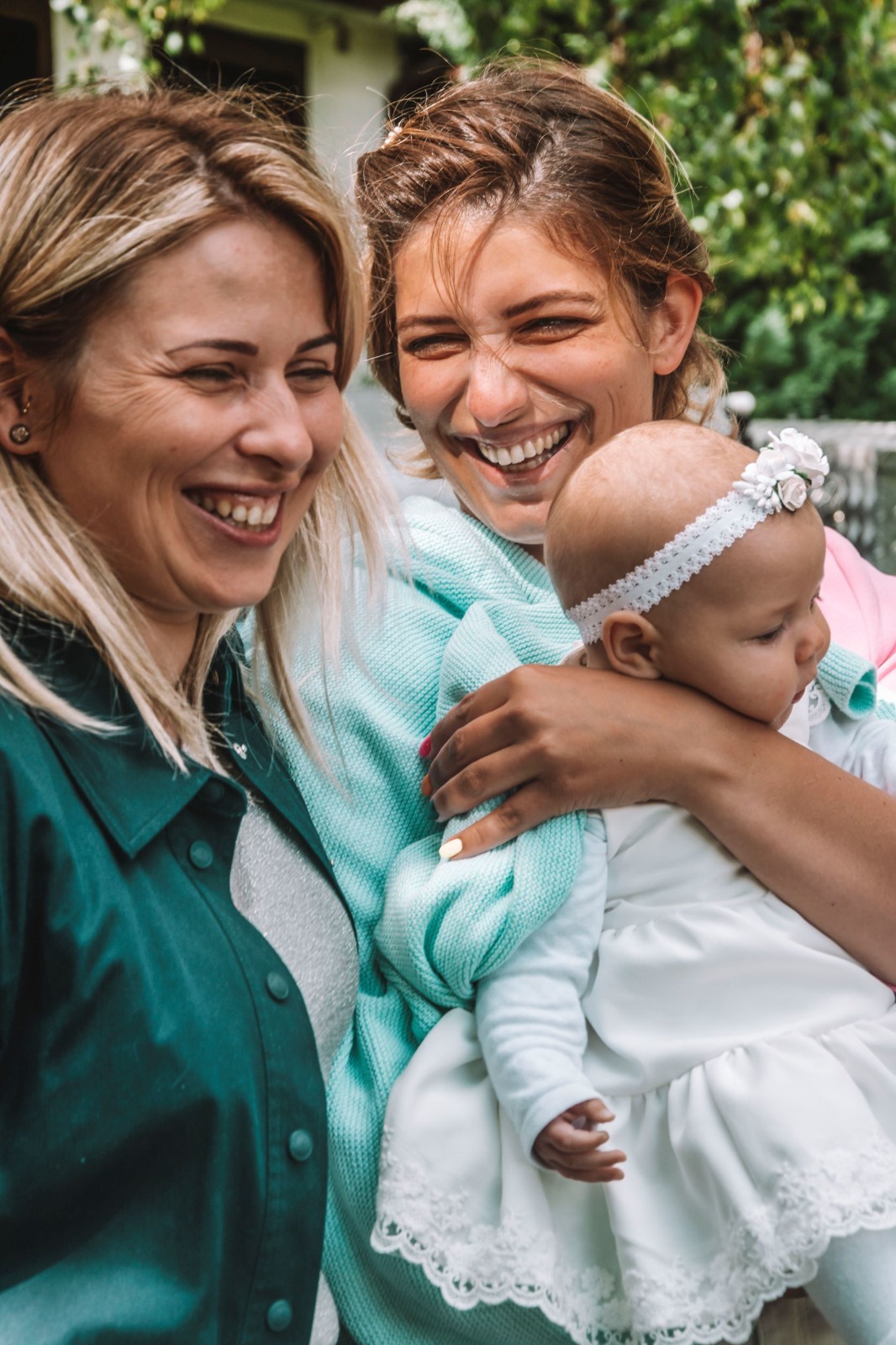 Dwie uśmiechnięte kobiety, jedna trzyma na rękach niemowlę w białej sukience z koronkowym wykończeniem i opaską z kwiatami na głowie. Widoczne kolorowe paznokcie kobiety trzymającej dziecko.