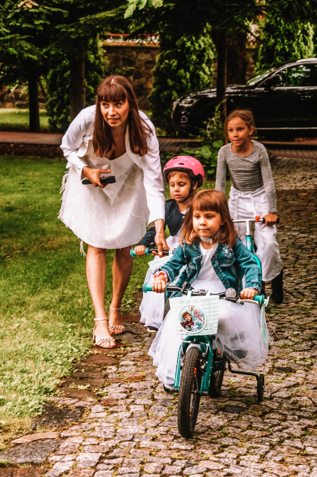 Kobieta w białej sukience i sandałach prowadzi grupę dziewczynek w białych sukienkach na rowerkach po kamiennej ścieżce w ogrodzie.