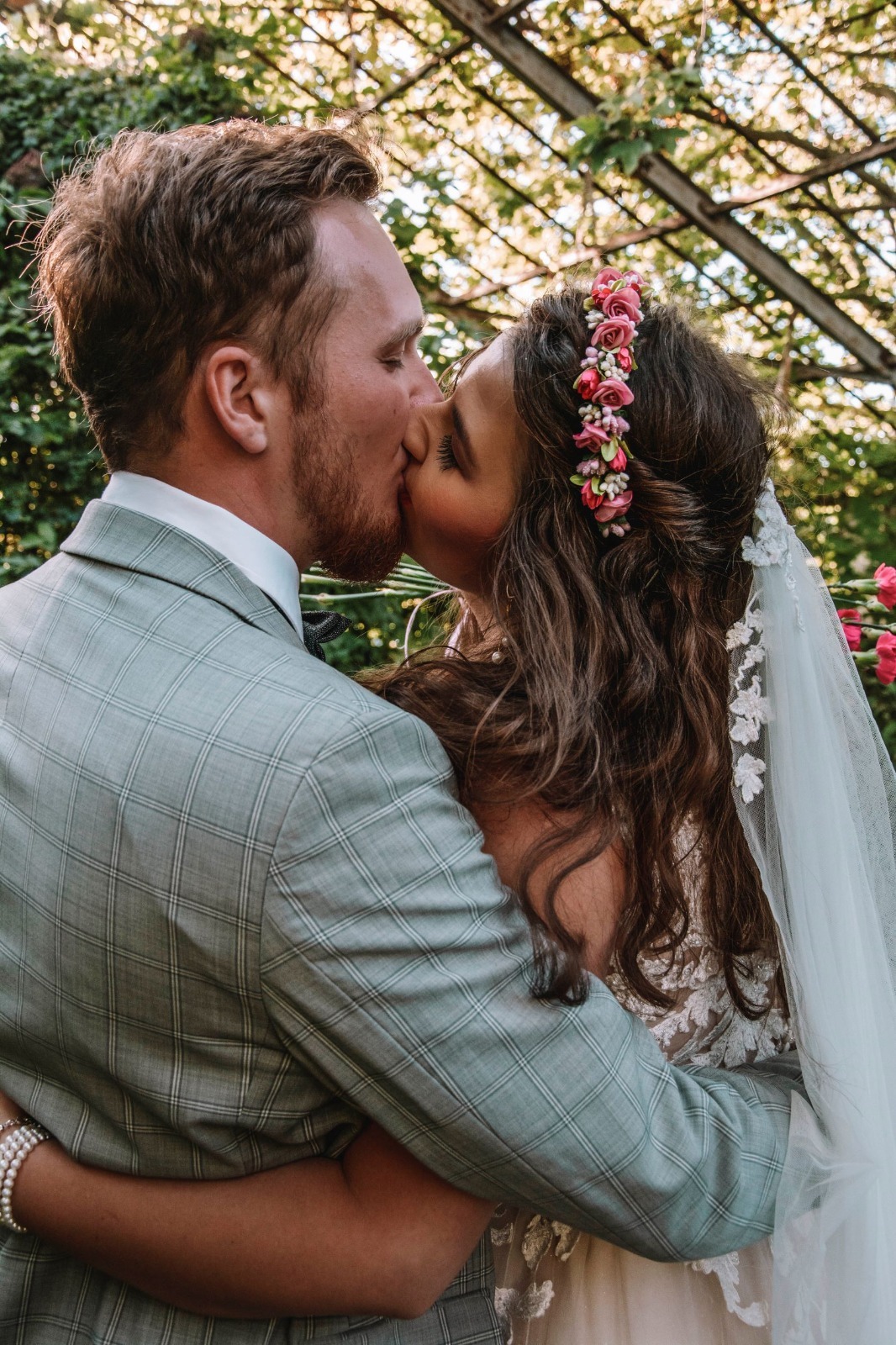 Pocałunek pary młodej pod konstrukcją z pnączy; panna młoda z wiankiem z różowych kwiatów i welonem z aplikacjami, pan młody w szarym garniturze w kratę.