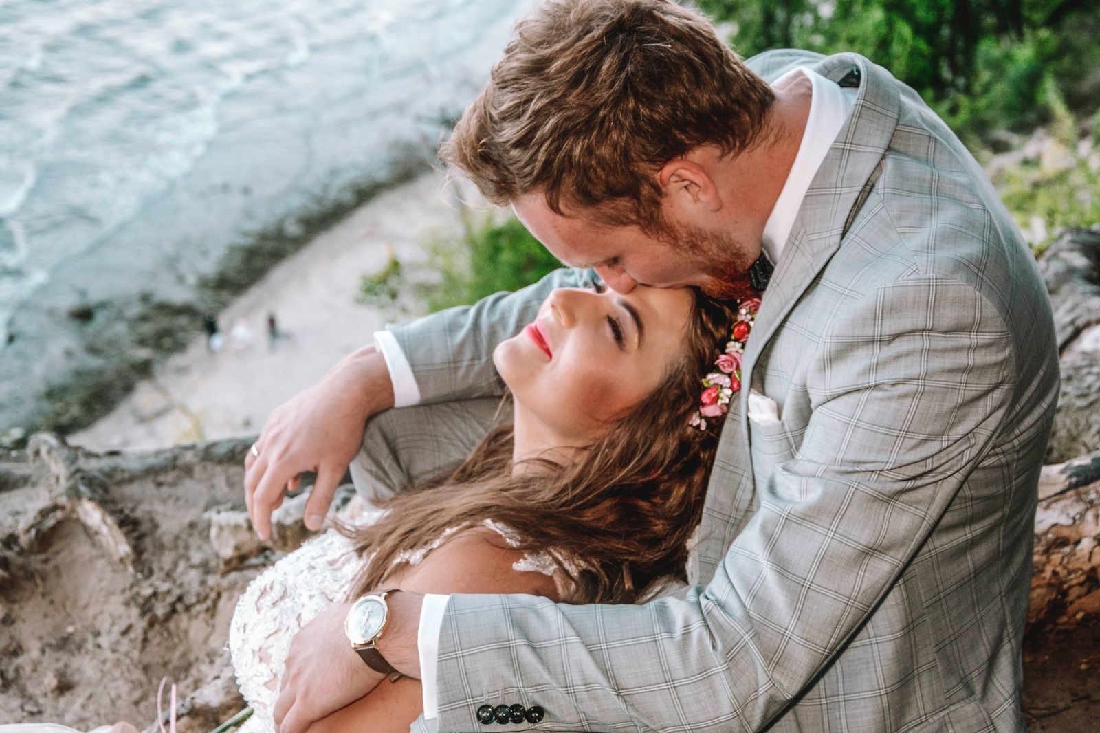 Para narzeczonych: mężczyzna w szarej kraciastej marynarce całuje kobietę w wianku z kwiatów na tle klifu i morza.