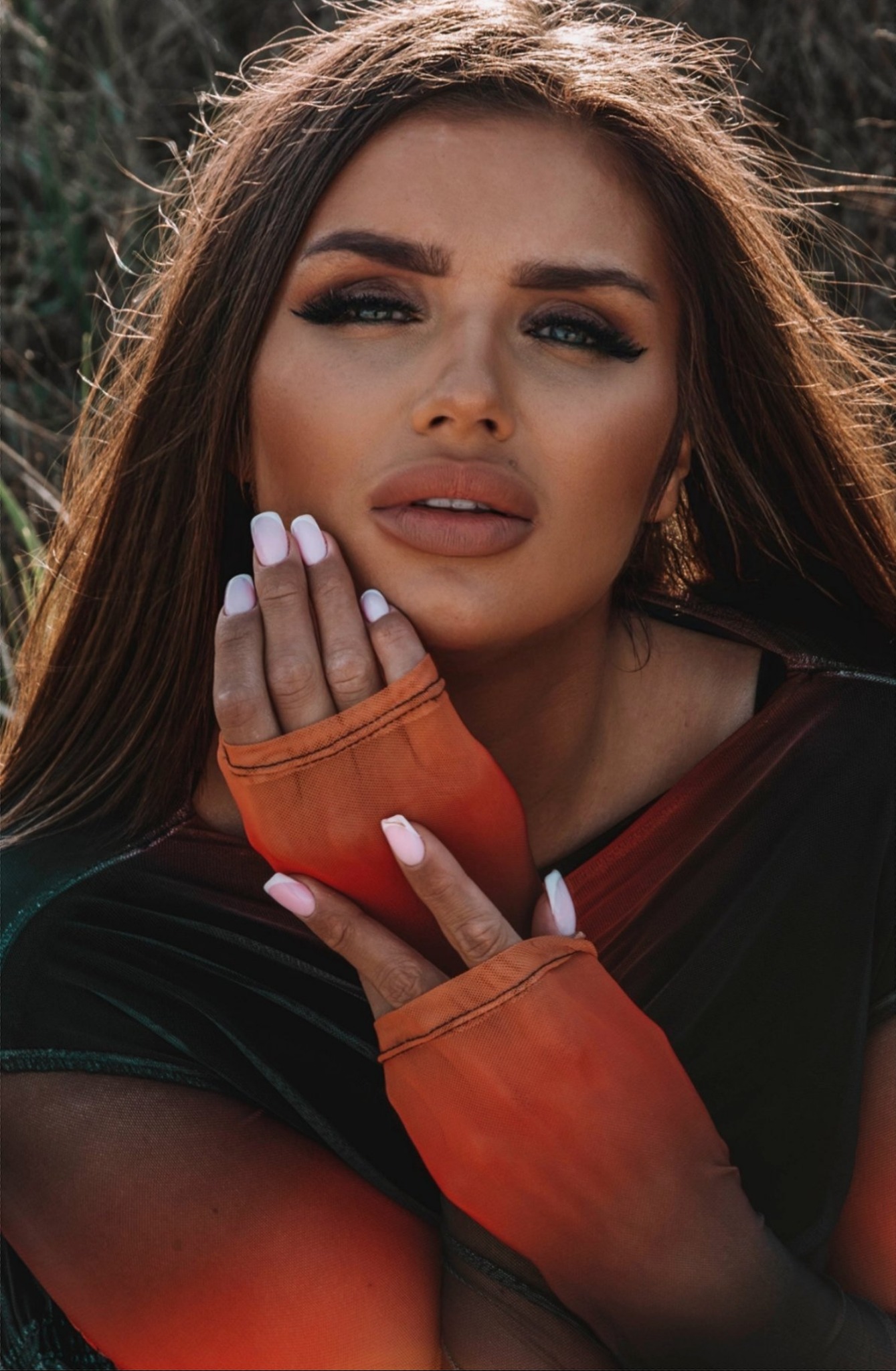 Portret kobiety z długimi brązowymi włosami, w pomarańczowych rękawiczkach z siateczki, delikatny makijaż, dłonie przy twarzy z manicure w stylu french.